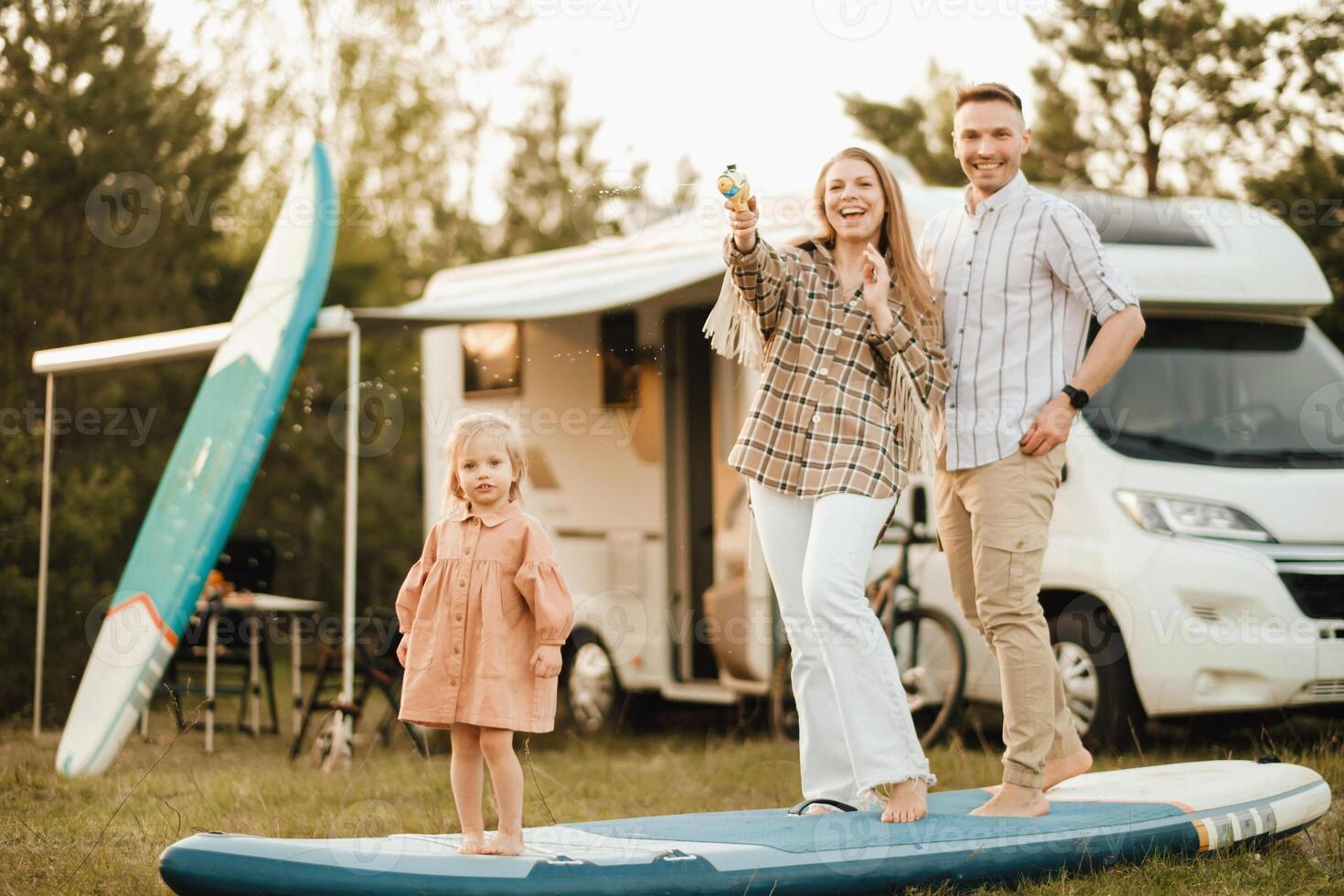 a família é em repouso Próximo para seus Móvel lar. pai, mãe e filha jogar em sup Pranchas com água pistolas perto a motorhome foto