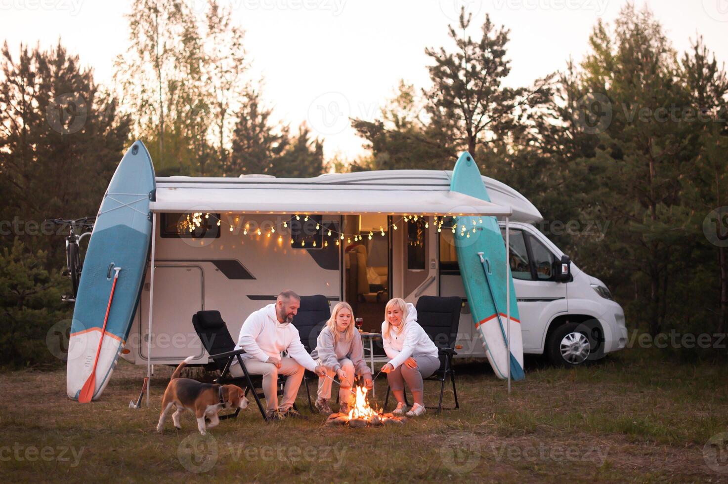 uma família cozinheiros salsichas em uma fogueira perto seus motorhome dentro a madeiras foto