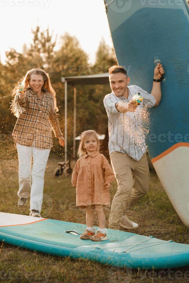 a família é em repouso Próximo para seus Móvel lar. pai, mãe e filha jogar em sup Pranchas com água pistolas perto a motorhome foto