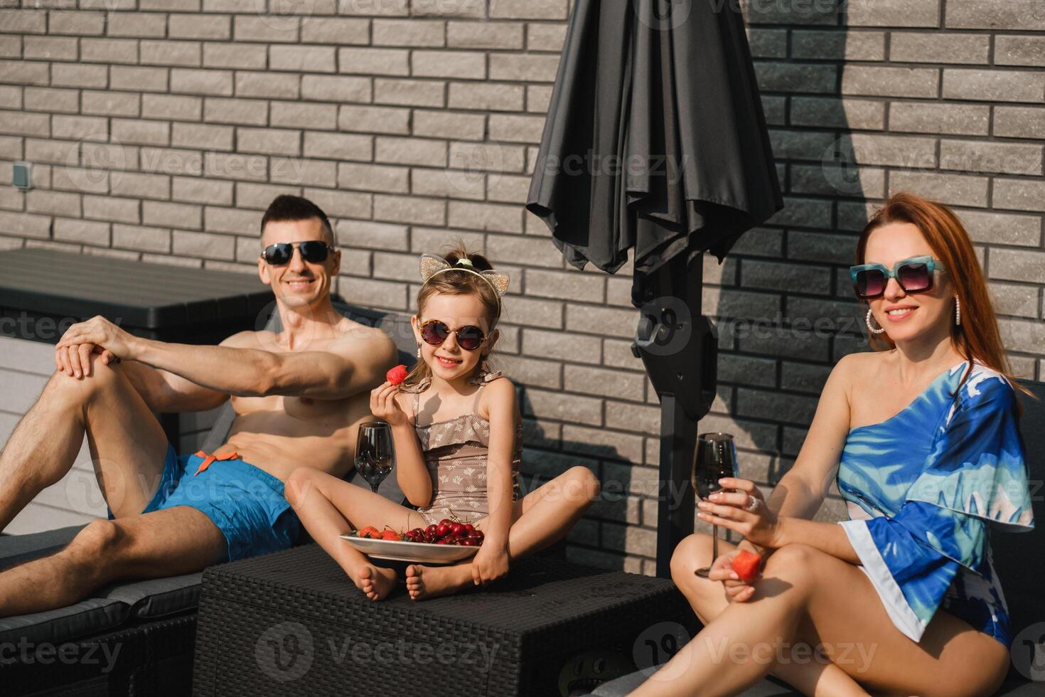 uma feliz família dentro maiôs tomar sol dentro verão em seus terraço em Sol camas foto