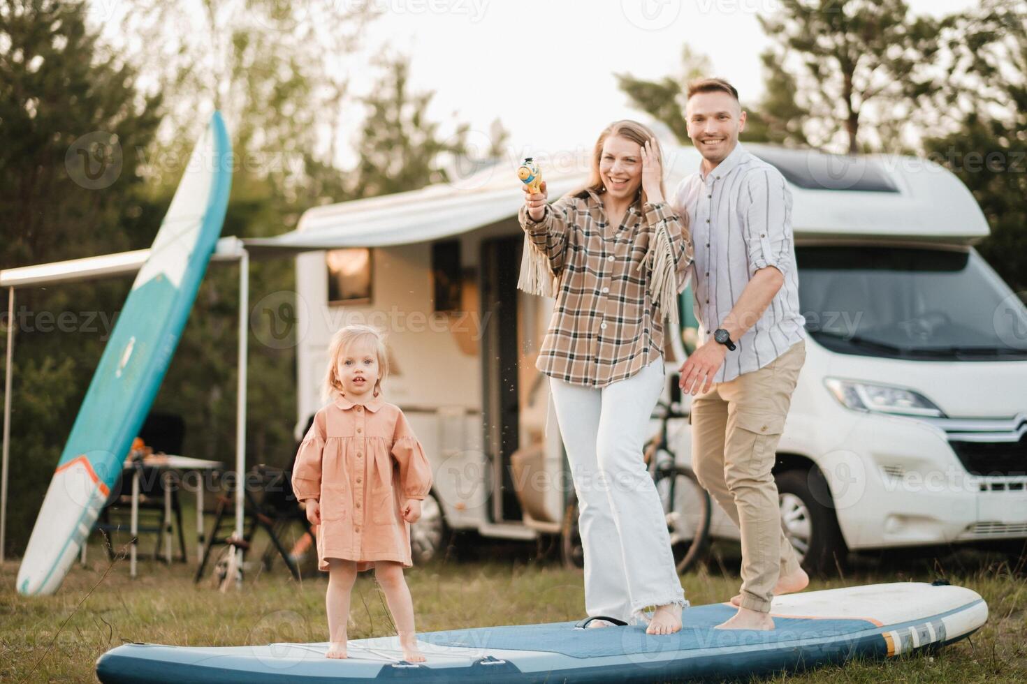 a família é em repouso Próximo para seus Móvel lar. pai, mãe e filha jogar em sup Pranchas com água pistolas perto a motorhome foto