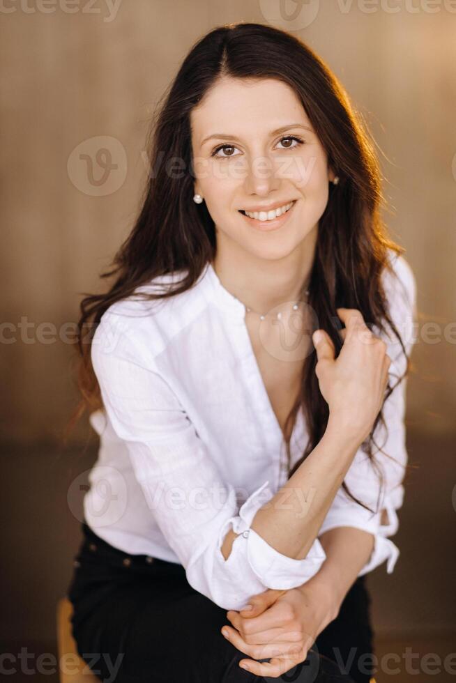 retrato do uma feliz jovem morena mulher dentro uma branco camisa sentado em uma cadeira foto