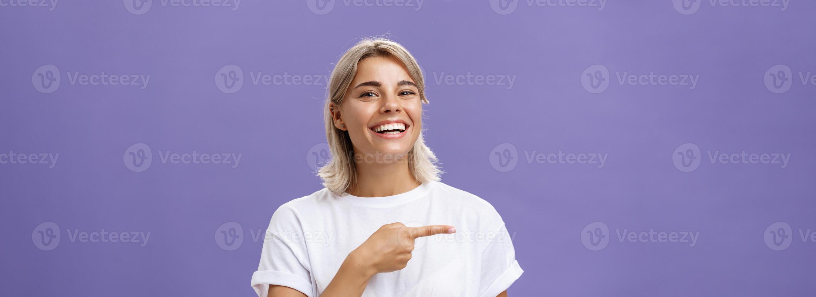 fechar-se tiro do divertido feliz e entretido boa aparência sociável mulher com justo cabelo e lindo sorrir sorrindo enquanto apontando esquerda com índice dedo mostrando impressionante cópia de espaço sobre roxa parede foto