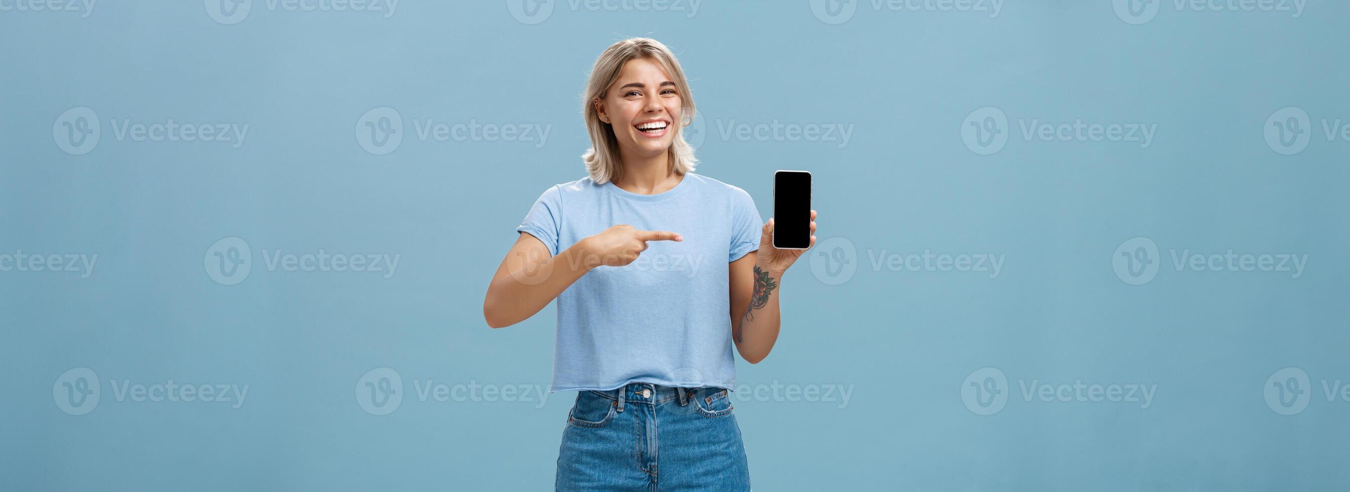 Veja às isto Hilário foto. entretido atraente feliz mulher com justo cabelo dentro casual camiseta e jeans calção mostrando Smartphone às Câmera apontando às dispositivo tela sorridente amplamente sobre azul parede foto