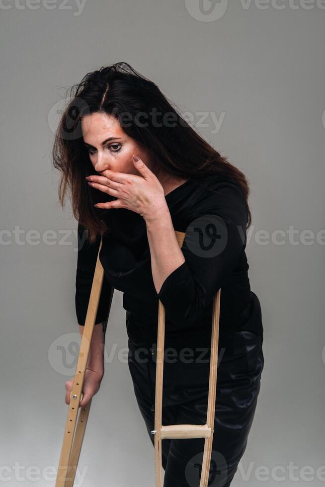 uma mulher espancado dentro Preto roupas com muletas dentro dela mãos em uma cinzento fundo foto
