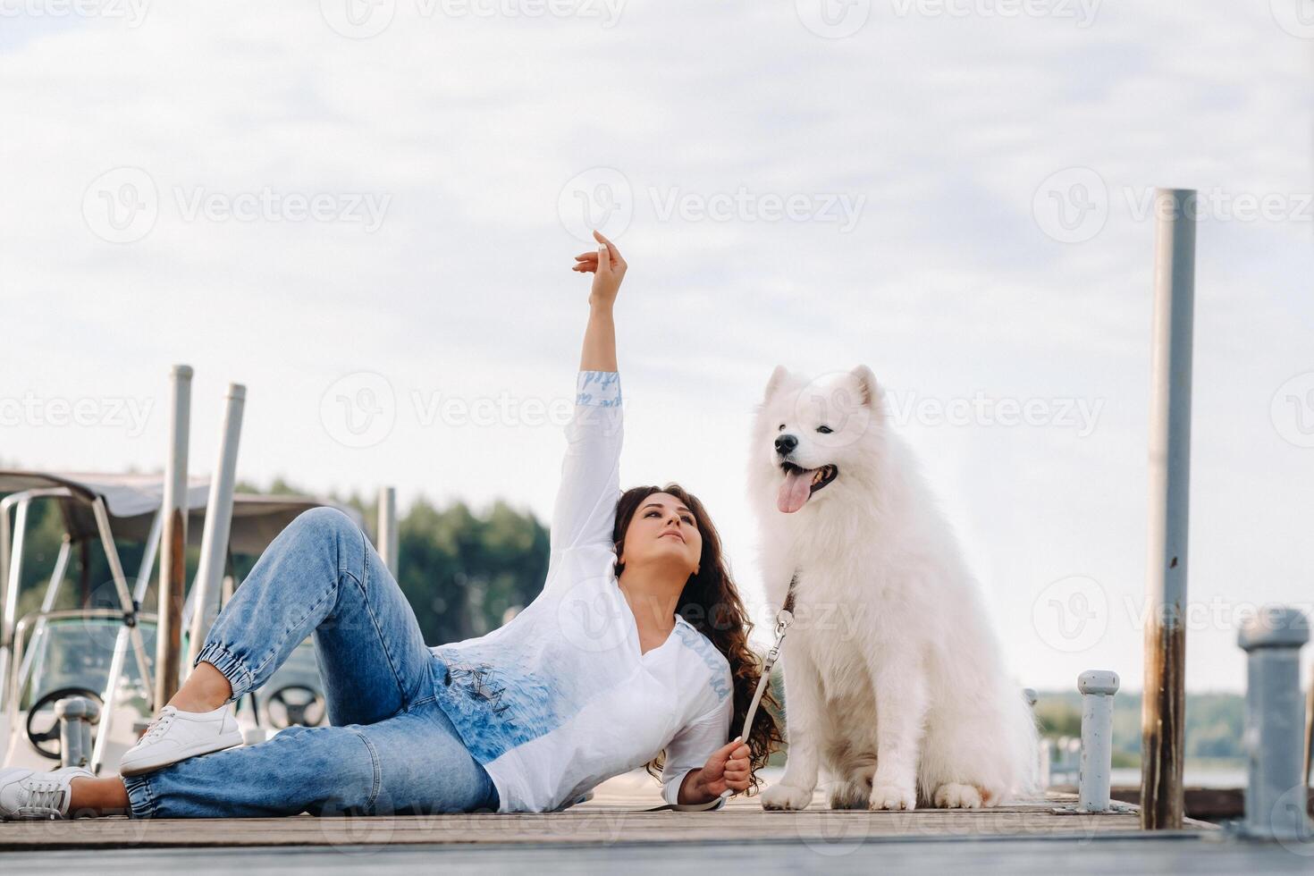 uma feliz mulher com uma grande branco cachorro mentiras em uma cais perto a mar às pôr do sol foto