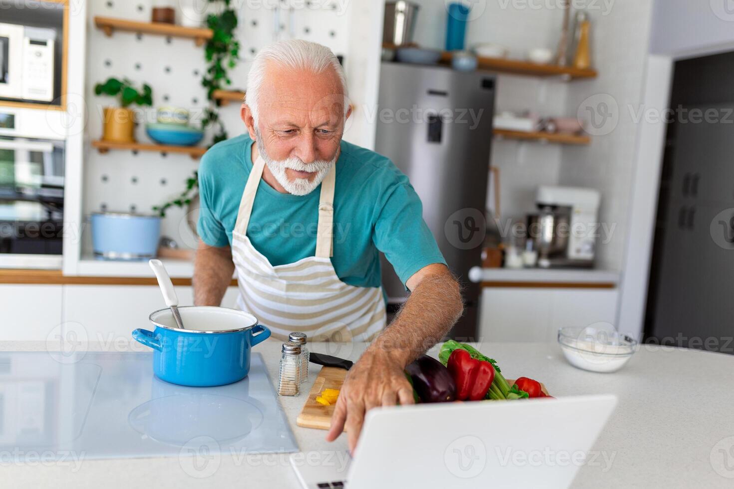 feliz Senior homem tendo Diversão cozinhando às casa - idosos pessoa preparando saudável almoço dentro moderno cozinha olhando às a recibo às dele computador portátil foto