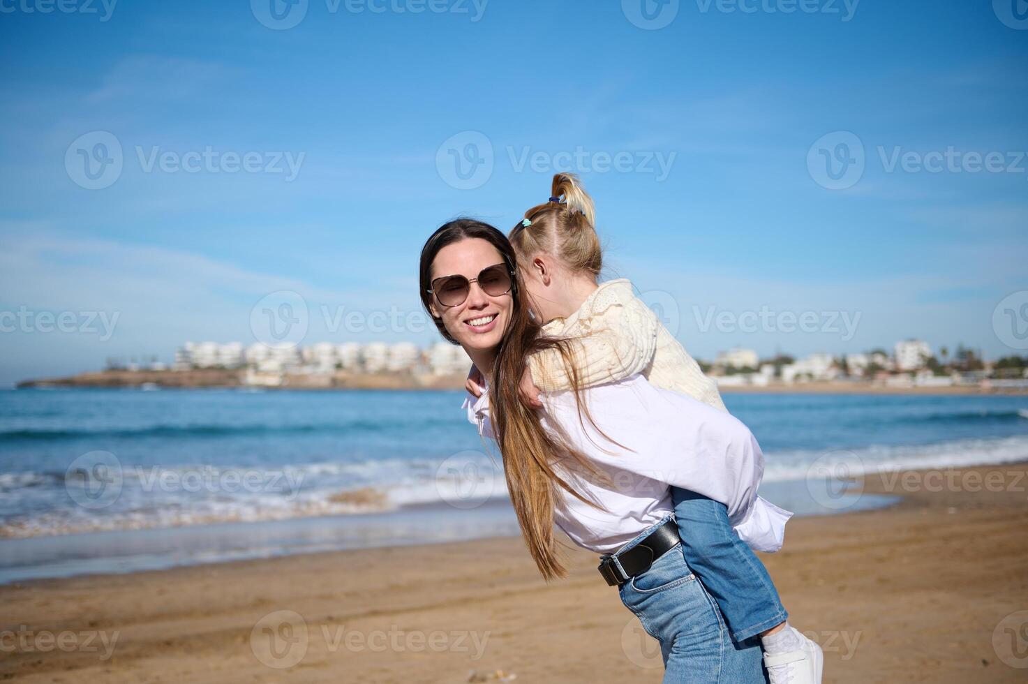 feliz família do uma jovem amoroso carinhoso mãe carregando pequeno filha em dela costas e caminhando em tropical arenoso de praia. foto