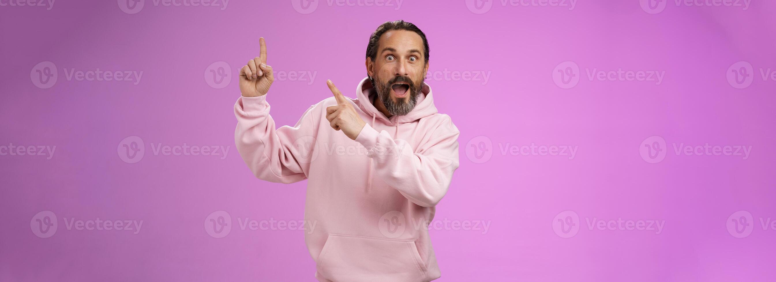 impressionado chocado animado legal hipster maduro decada de 50 homem barbudo cinzento cabelo dentro Rosa moletom com capuz recontar incrível história apontando acima índice dedos ampliar olhos surpreso emocionados impressionante propaganda foto