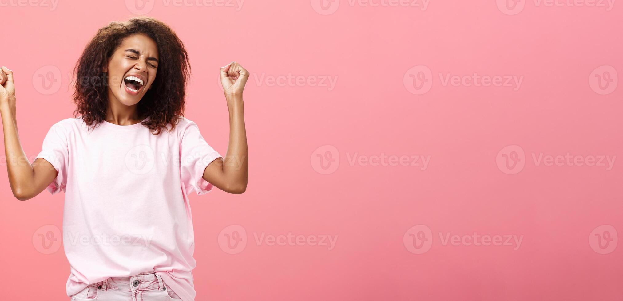 espantado e feliz triunfante africano americano desportista a comemorar vitória gritando a partir de impressionante emocionante sentimentos fechamento olhos levantando punhos altura dentro ganhar gesto em pé sobre Rosa fundo foto