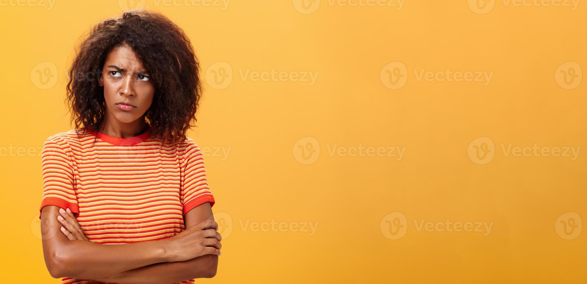 porque vida tão cruel e injusto. sombrio descontente e triste fofa africano americano jovem mulher com encaracolado Penteado cruzando braços em peito dentro ofendido pose franzindo a testa olhando esquerda com ciumes e infeliz foto