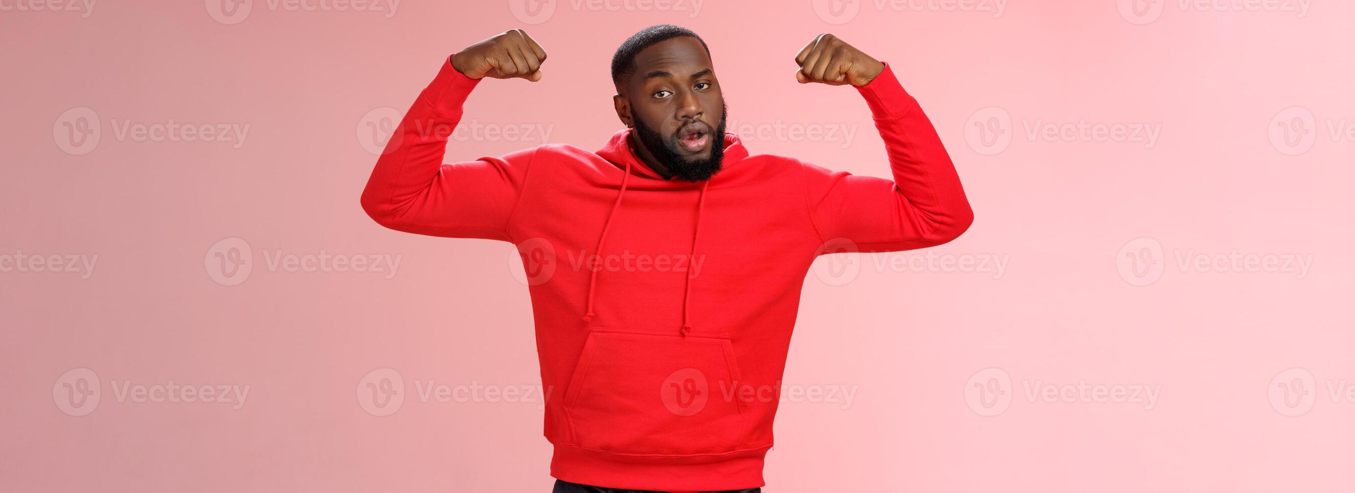 Forte atrevido sério africano americano desportista dentro vermelho moletom com capuz se gabar exibir levantar mãos exercício mostrar grande músculos bíceps, em pé confiante Rosa fundo foto