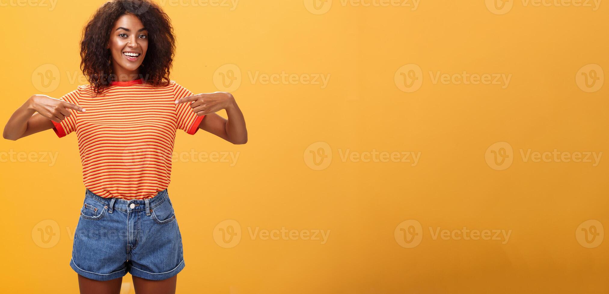 Ei escolher mim Eu seu menina olhando para. retrato do encantador de aparência amigável ambicioso pele escura fêmea com afro Penteado apontando às peito orgulhosamente e alegre posando contra laranja fundo foto