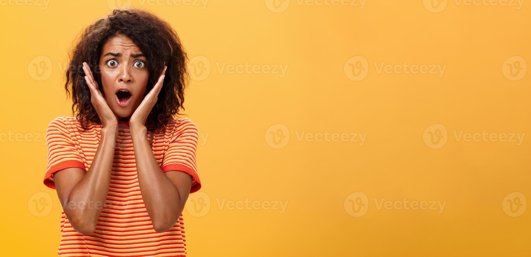 cintura para cima tiro do chocado preocupado em pânico afro-americano mulher com afro Penteado dentro na moda camiseta ofegante segurando Palmeiras em face a partir de surpresa posando perturbado contra laranja parede foto