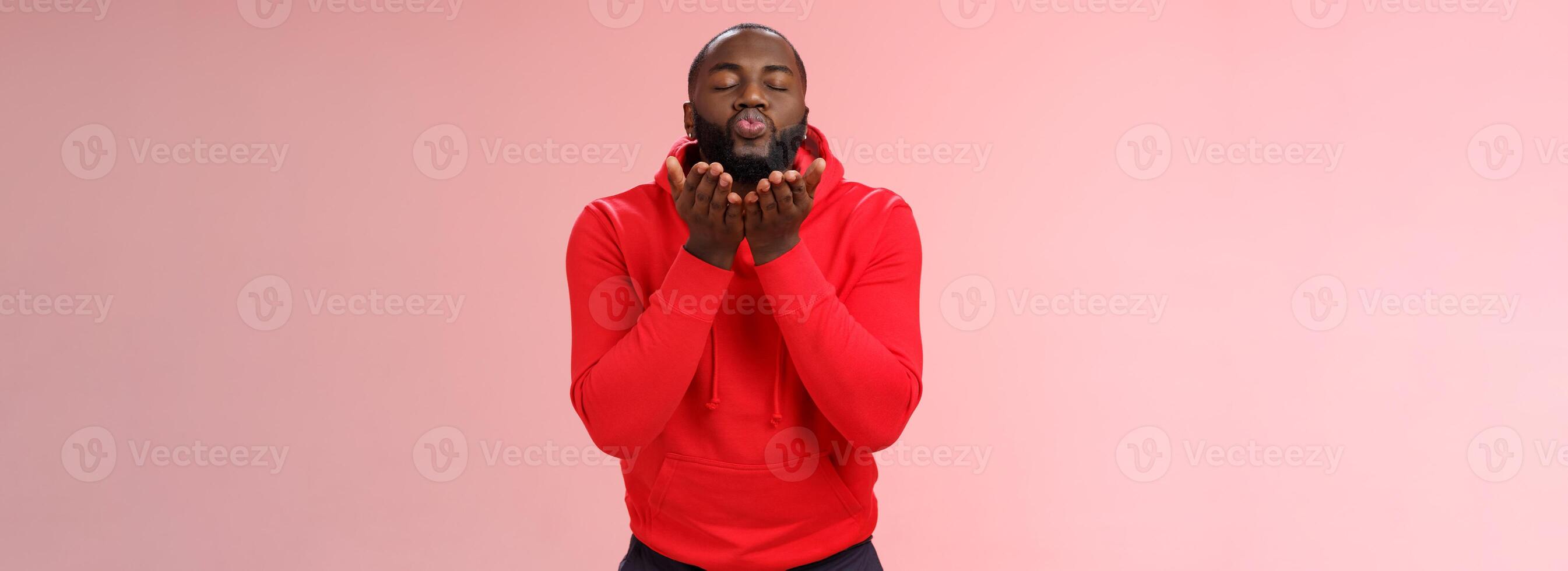 encantador fofa africano americano barbudo cara dentro vermelho moletom com capuz fechar olhos dobrando lábios aguarde Palmeiras perto boca enviando apaixonado ar beijo sopro mwah Câmera, em pé adorável Rosa fundo foto