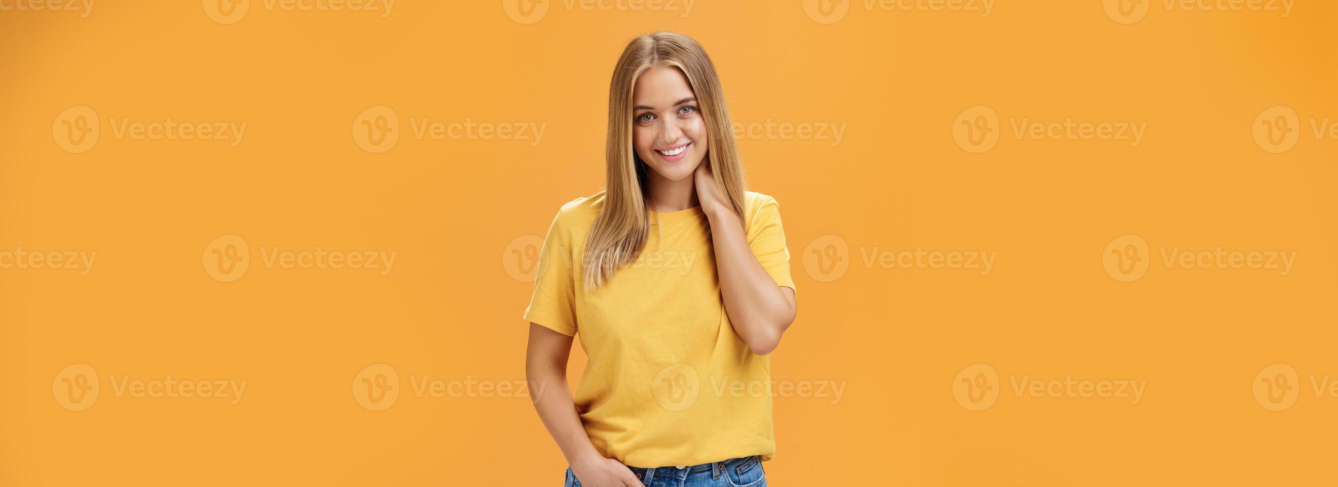 retrato do tímido e tímido feminino menina com bronzeado e em linha reta fait cabelo fricção pescoço e sorridente sensualmente com feliz despreocupado expressão segurando mão dentro bolso posando contra laranja fundo foto