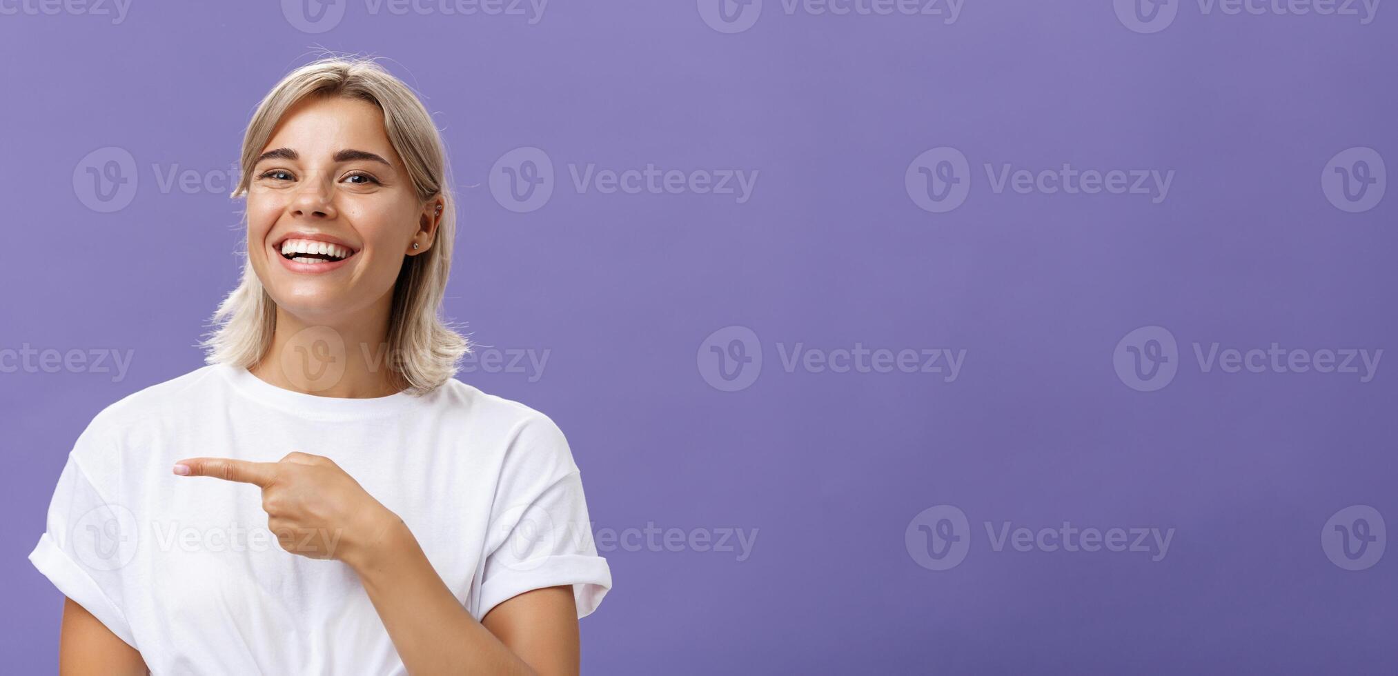 fechar-se tiro do divertido feliz e entretido boa aparência sociável mulher com justo cabelo e lindo sorrir sorrindo enquanto apontando esquerda com índice dedo mostrando impressionante cópia de espaço sobre roxa parede foto