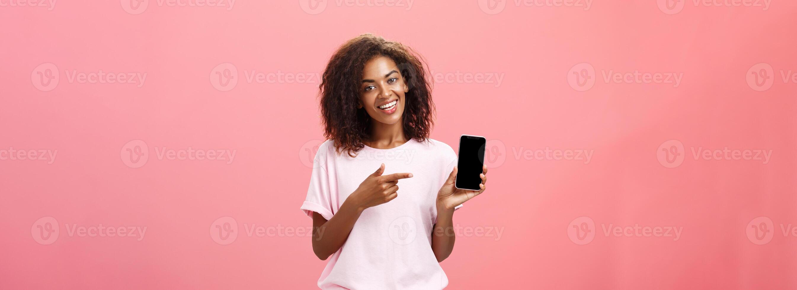 sugerir isto modelo do telefone. feliz encantador africano americano cabelo encaracolado mulher dentro à moda equipamento segurando espertinho e apontando às dispositivo tela enquanto recomendando legal aplicativo sobre Rosa parede foto