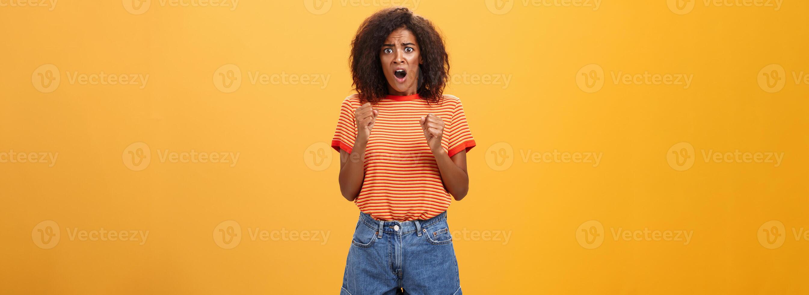 mulher sobre para pânico audição chocante preocupado notícias. retrato do intenso nervoso e ansioso pele escura adulto menina com afro Penteado aperto punhos abertura boca olhando preocupado sobre laranja parede foto