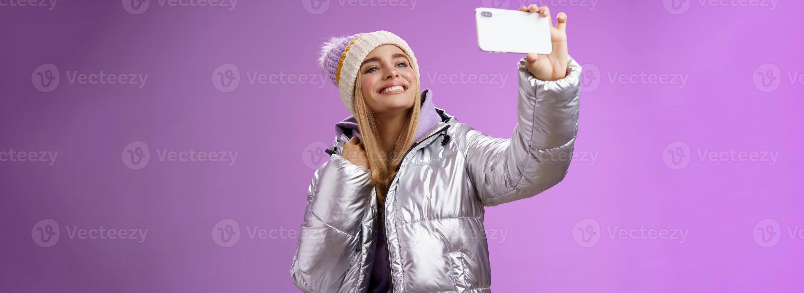 feliz Deleitado despreocupado louro encantador europeu mulher dentro prata inverno Jaqueta chapéu levantando Smartphone horizontalmente levando selfie sorridente Móvel telefone mostrar, roxa fundo foto