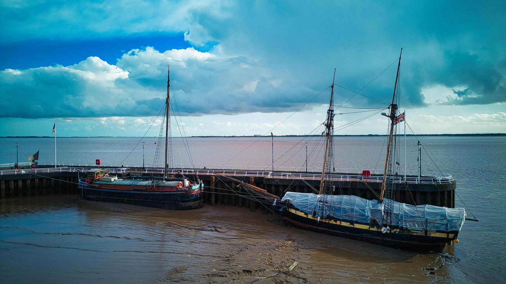 vintage Navegando navios ancorado às uma de madeira cais debaixo uma nublado céu, com calma águas e turvar litoral dentro casco, Inglaterra. foto