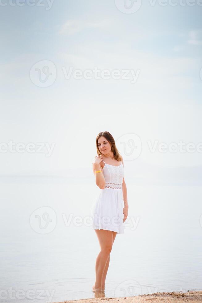 lindo mulher dentro uma branco vestir caminhando em a praia.relaxado mulher respiração fresco ar, emocional sensual mulher perto a mar, desfrutando verão.viagem e período de férias. liberdade e inspiração conceito foto