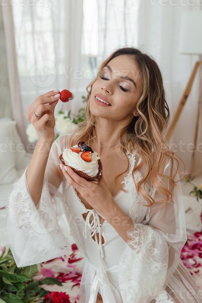 uma lindo menina é comendo uma morango shortcake às lar, cercado de Primavera flores em uma ampla cama. uma quarto decorado com buquês do peônias. foto