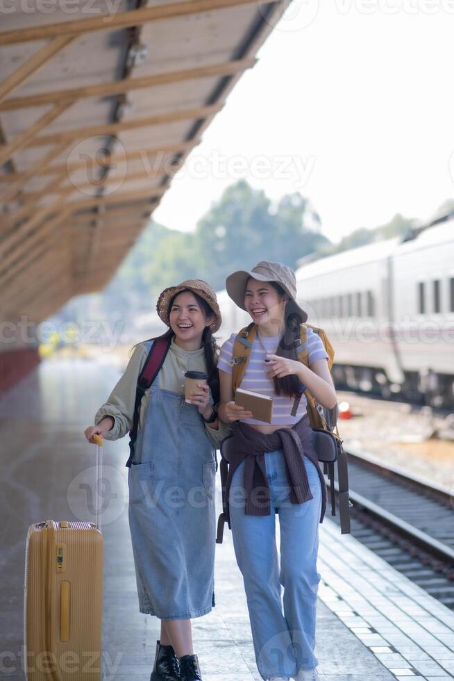 dois jovem ásia amigos meninas com mochilas às estrada de ferro estação esperando para trem, dois lindo mulheres caminhando ao longo plataforma às trem estação foto