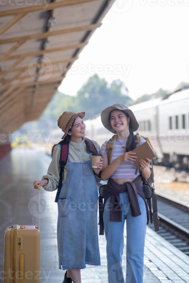 dois jovem ásia amigos meninas com mochilas às estrada de ferro estação esperando para trem, dois lindo mulheres caminhando ao longo plataforma às trem estação foto