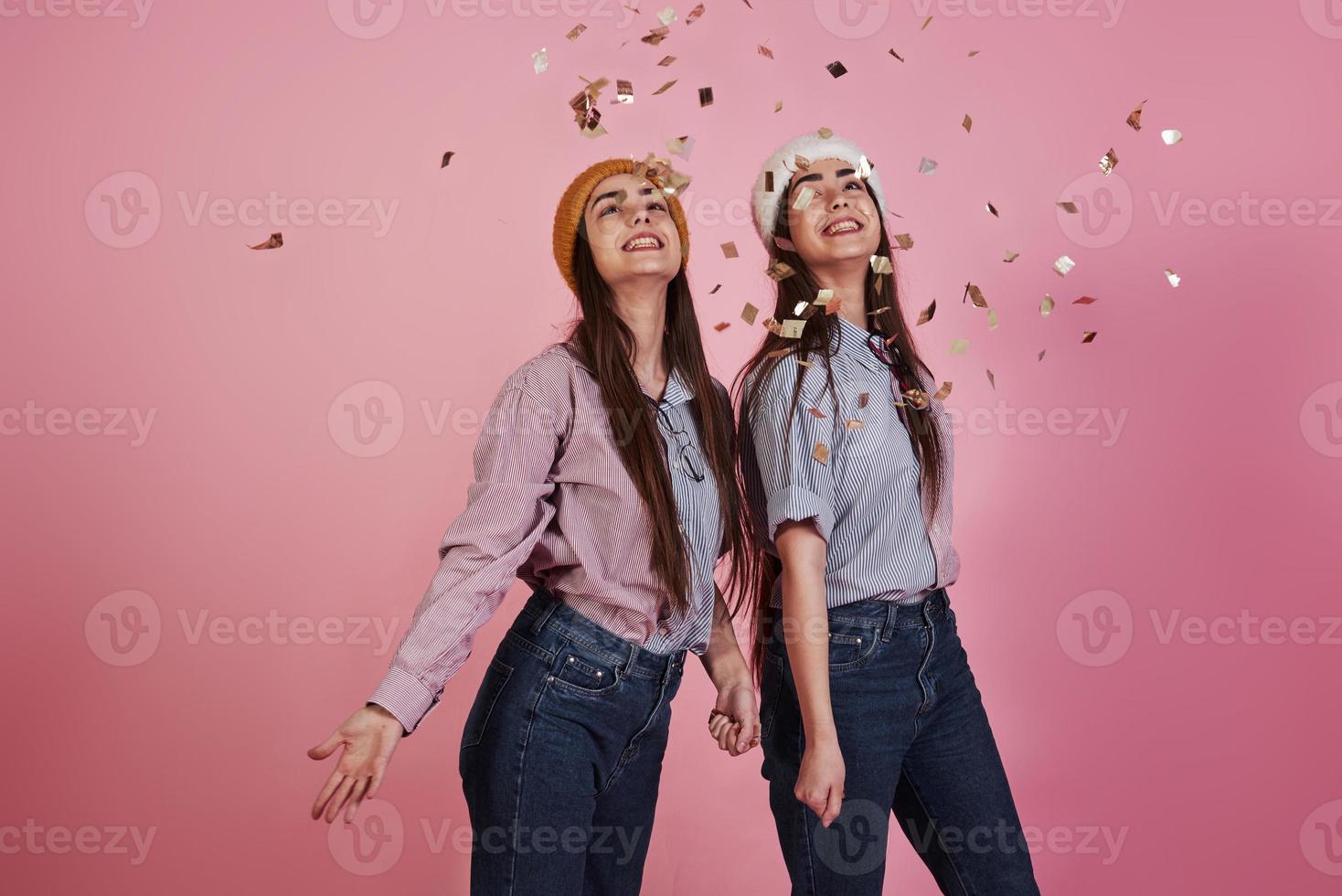 humor de feriado. concepção de ano novo. dois gêmeos jogando confetes dourados para o alto no estúdio com fundo rosa foto