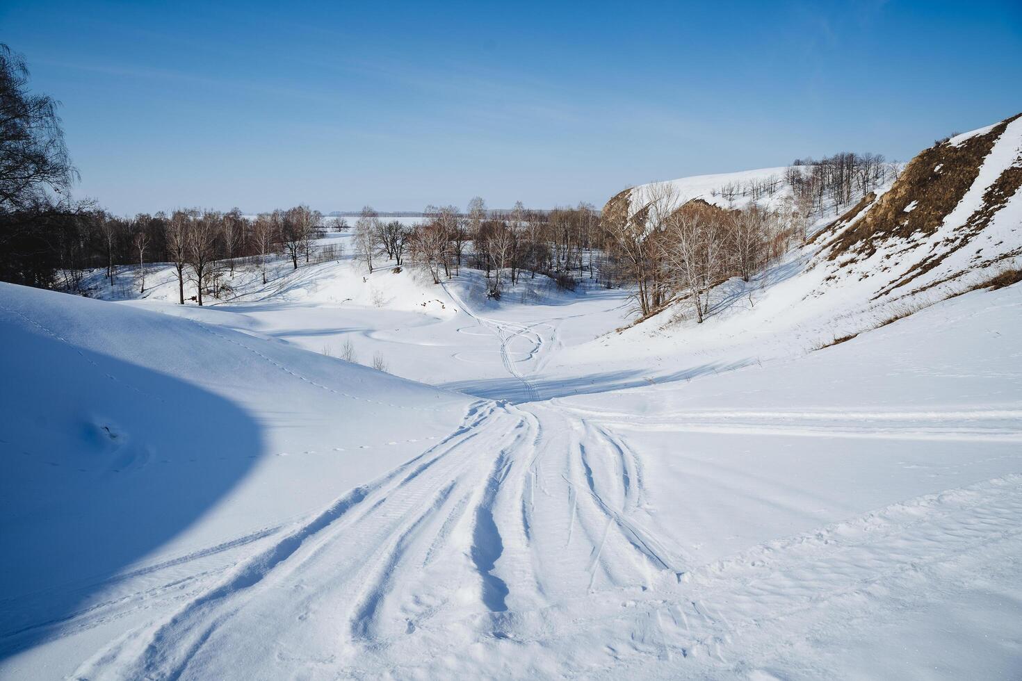 moto de neve faixas em branco neve, a estrada passes através uma coberto de neve rio, inverno paisagem, ensolarado clima Claro céu, inverno frio foto