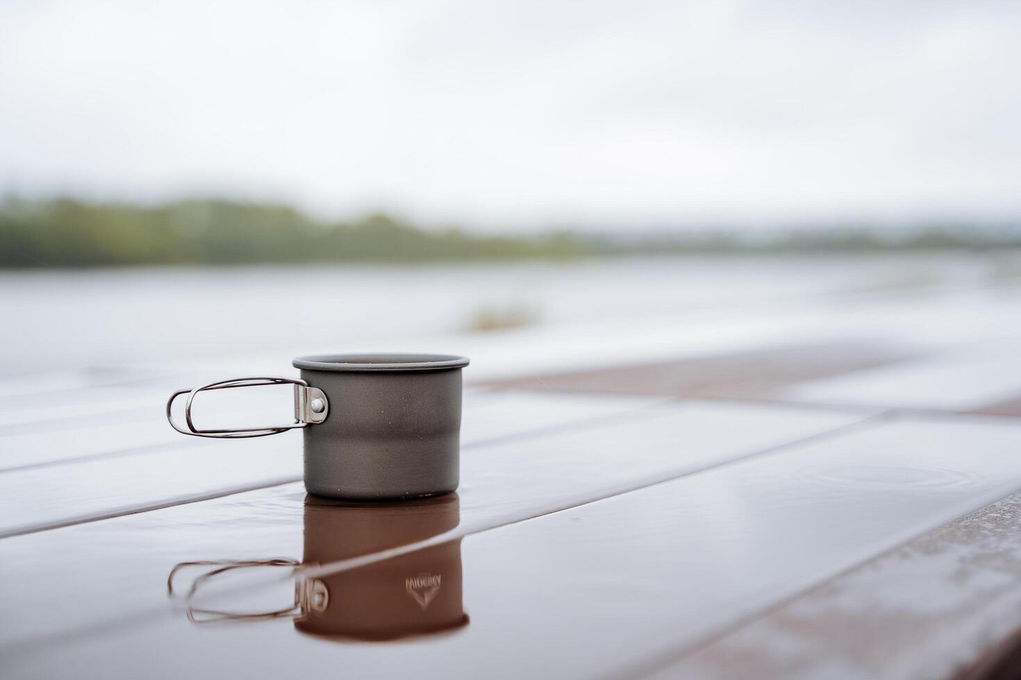 alumínio caneca carrinhos em a mesa dentro uma poça do água, reflexão dentro a água, chá vidro, café caneca caminhada, viagem para natureza, acampamento Comida beber chá foto