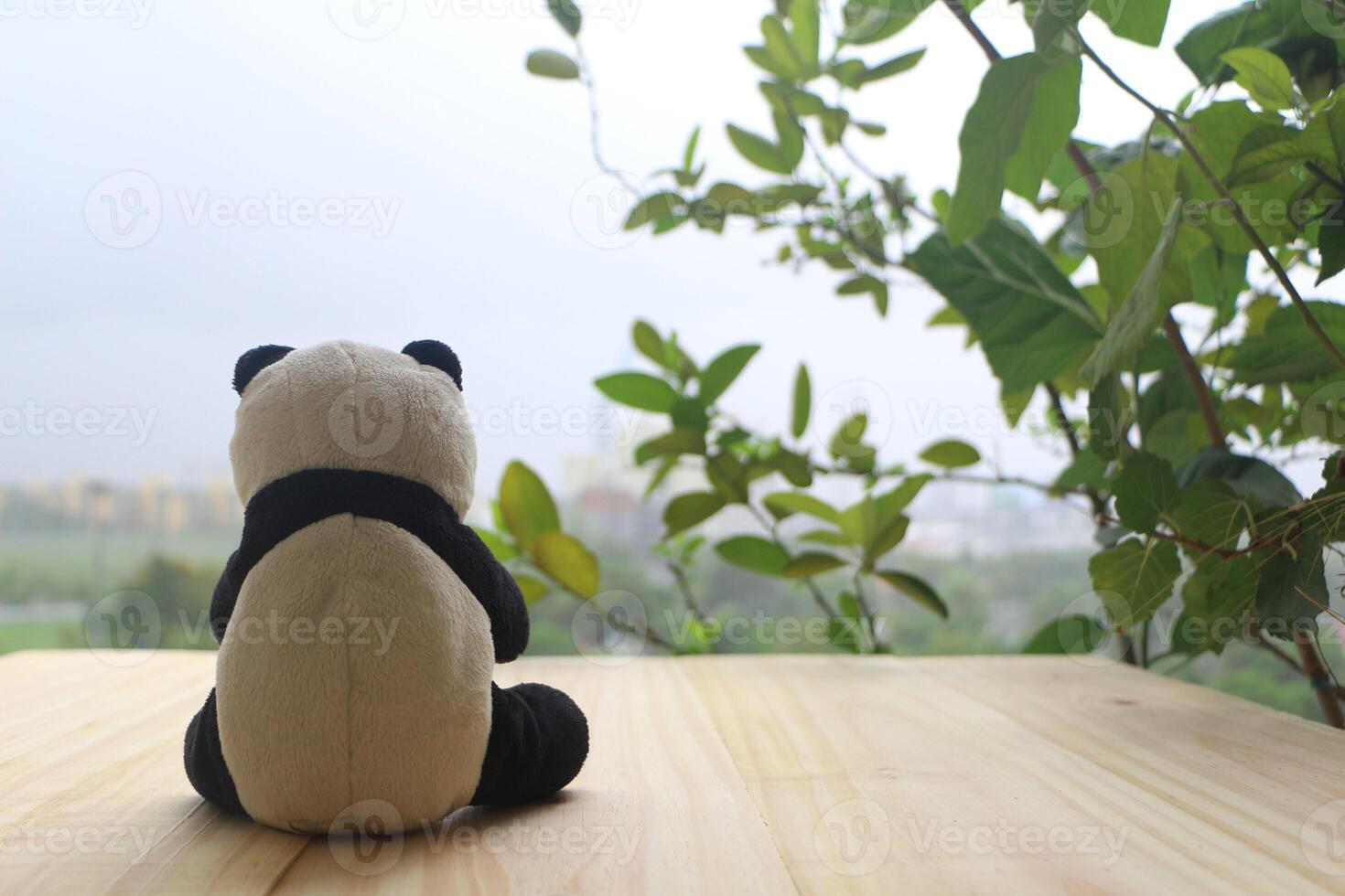 Preto e branco panda boneca sentado sozinho em a de madeira sacada, olhando Fora para a paisagens urbanas. foto