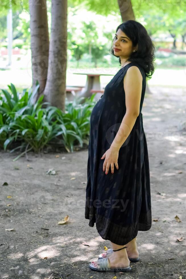 uma grávida indiano senhora poses para ao ar livre gravidez tiro e mãos em barriga, indiano grávida mulher coloca dela mão em dela estômago com uma maternidade vestir às sociedade parque, grávida lado de fora maternidade tiro foto