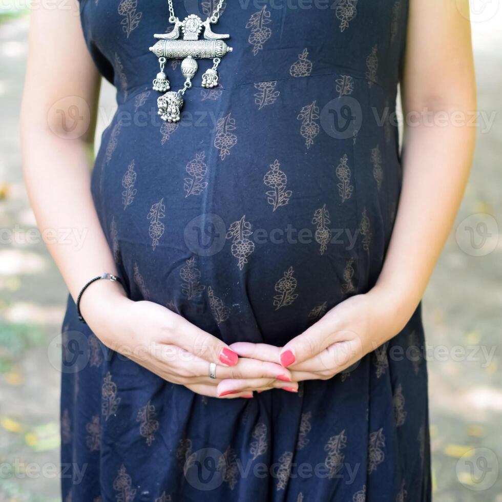 uma grávida indiano senhora poses para ao ar livre gravidez tiro e mãos em barriga, indiano grávida mulher coloca dela mão em dela estômago com uma maternidade vestir às sociedade parque, grávida lado de fora maternidade tiro foto