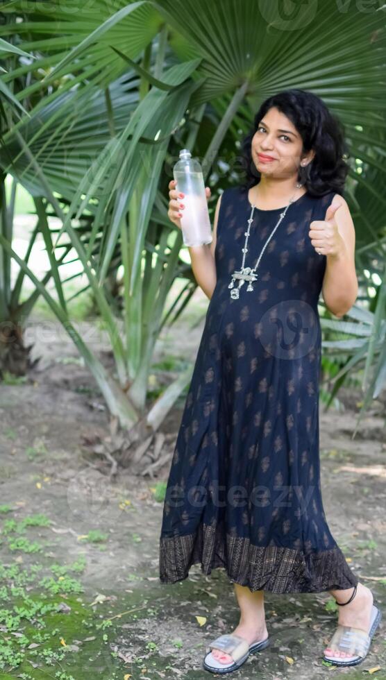 uma grávida indiano senhora poses para ao ar livre gravidez tiro e mãos em barriga, indiano grávida mulher coloca dela mão em dela estômago com uma maternidade vestir às sociedade parque, grávida lado de fora maternidade tiro foto