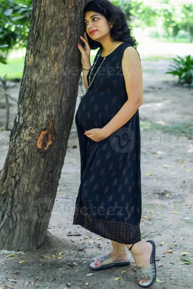 uma grávida indiano senhora poses para ao ar livre gravidez tiro e mãos em barriga, indiano grávida mulher coloca dela mão em dela estômago com uma maternidade vestir às sociedade parque, grávida lado de fora maternidade tiro foto