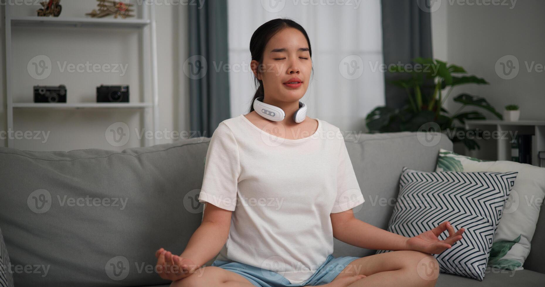 feliz jovem mulher vestem automático massagem pescoço meditando fazendo uma lótus pose às casa com olhos fechado, relaxante corpo e mente sozinho em sofá dentro a vivo quarto, relaxamento estilo de vida foto