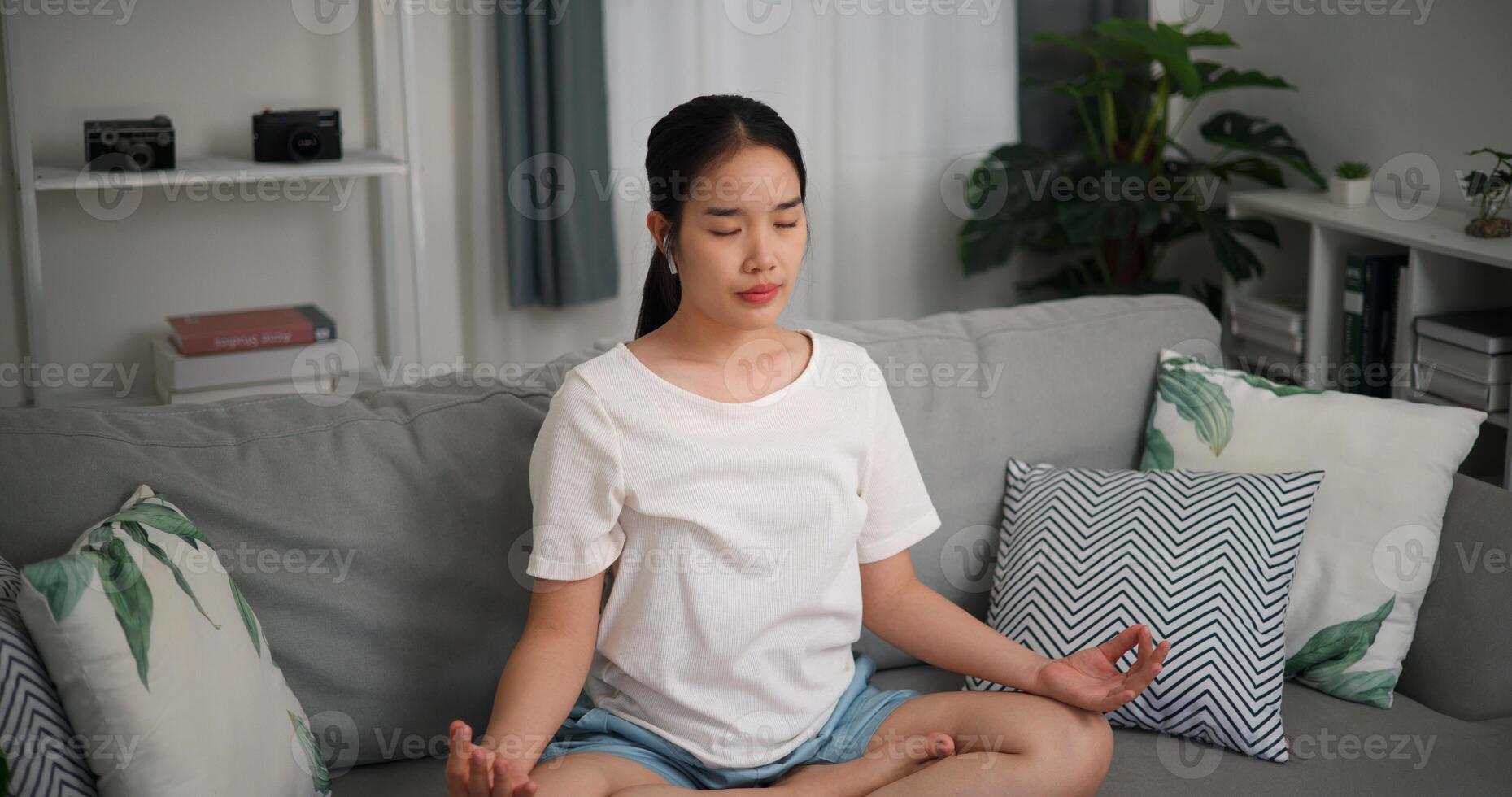 Alto ângulo visualizar, feliz jovem mulher vestem sem fio fones de ouvido meditando fazendo uma lótus pose às casa com olhos fechado, relaxante corpo e mente sozinho em sofá dentro a vivo quarto, relaxamento estilo de vida foto