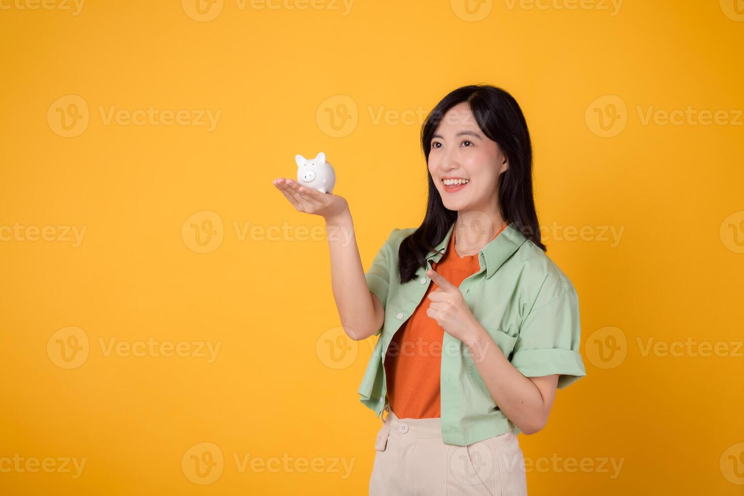 alegre jovem ásia mulher dentro dela 30s, vestindo laranja camisa e verde saltador, exibindo porquinho banco enquanto apontando dedo para livre cópia de espaço em amarelo fundo. financeiro dinheiro conceito. foto