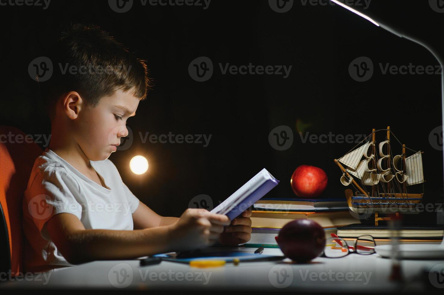 concentrado estudante lendo livro às mesa com livros, plantar, lâmpada, cor lápis, maçã, e livro didático foto
