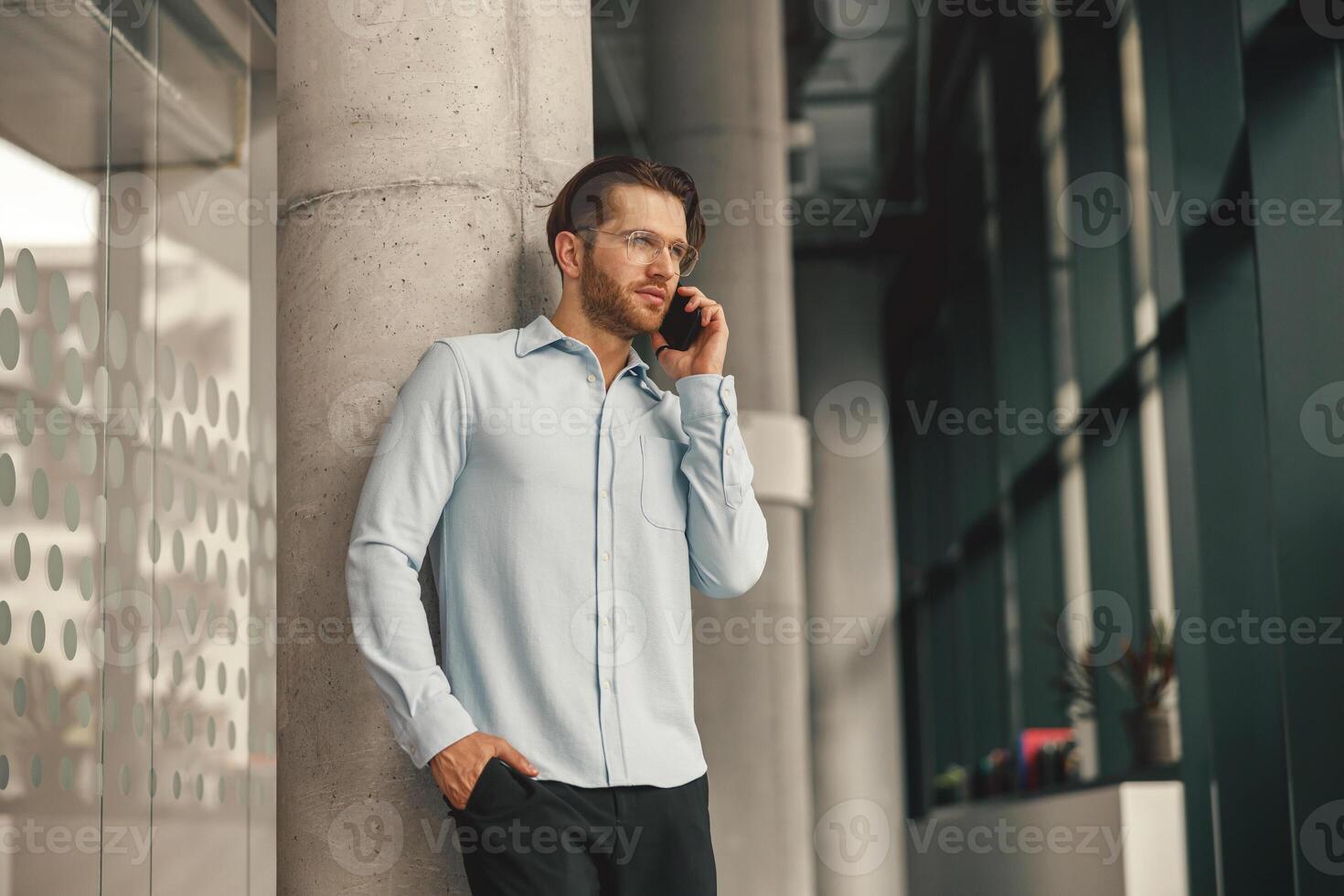 focado masculino empreendedor falando telefone com cliente enquanto em pé dentro escritório e olhando às lado foto