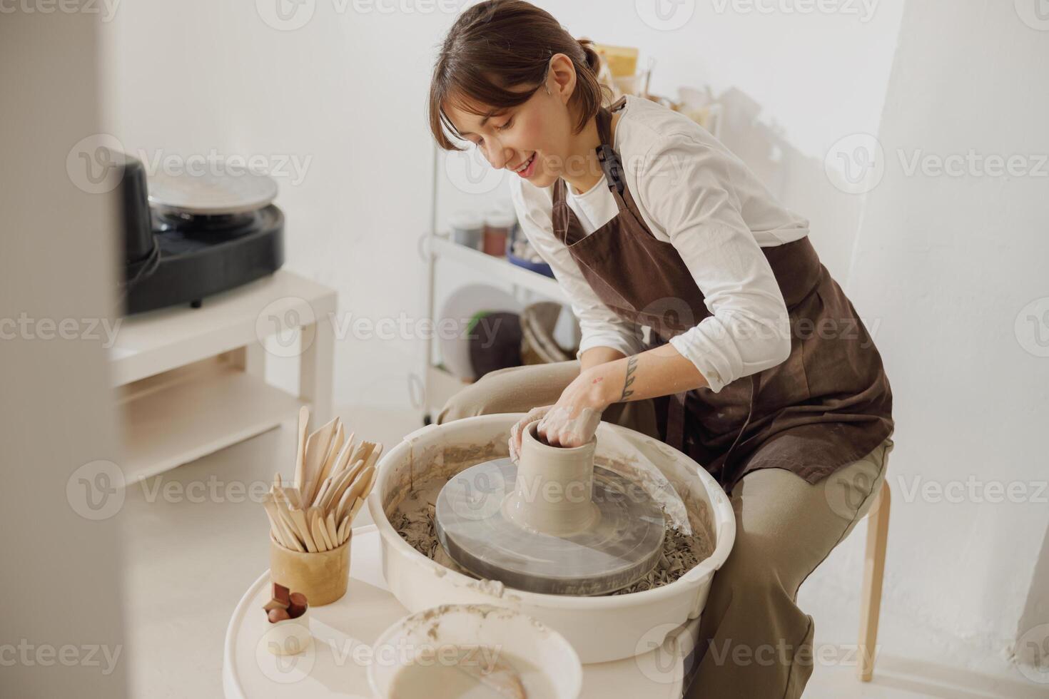 sorridente fêmea artesão dentro avental sentado em Banco com cerâmica roda e fazer argila Panela foto