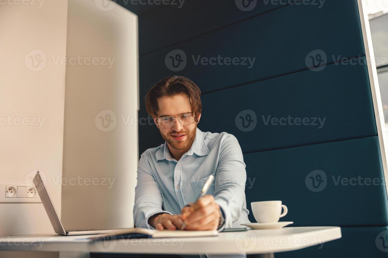 sorridente masculino trabalhador autonomo fazer notas durante trabalhando em computador portátil sentar escrivaninha dentro coworking foto