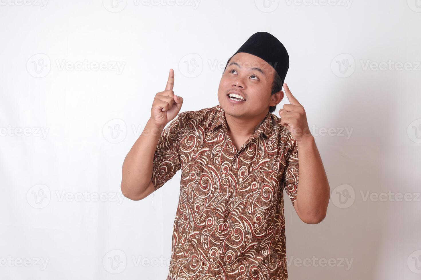 retrato do animado ásia homem vestindo batik camisa e Songkok sorridente e olhando às a Câmera apontando com dois mãos e dedos para acima. isolado imagem em cinzento fundo foto