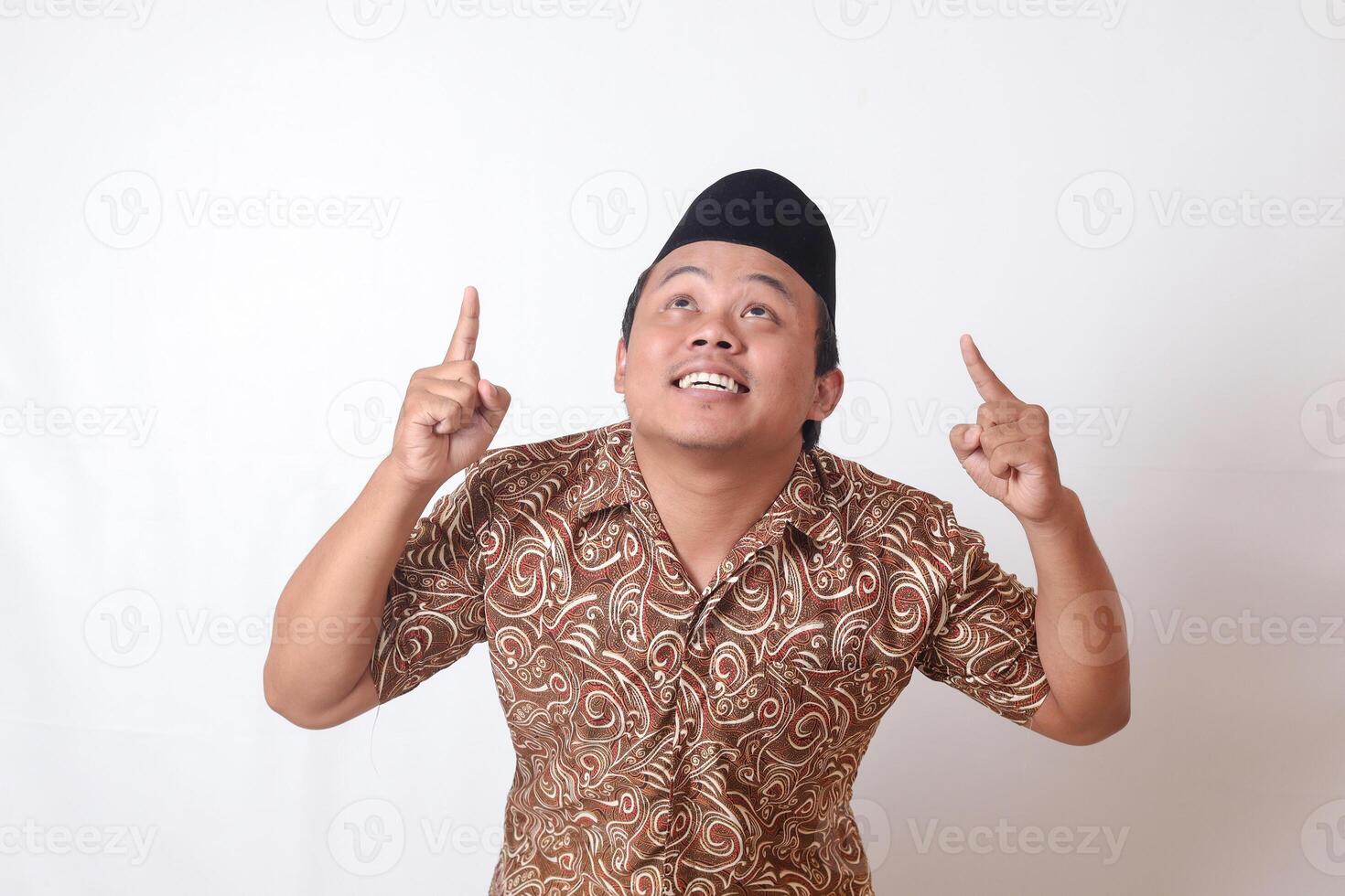 retrato do animado ásia homem vestindo batik camisa e Songkok sorridente e olhando às a Câmera apontando com dois mãos e dedos para acima. isolado imagem em cinzento fundo foto