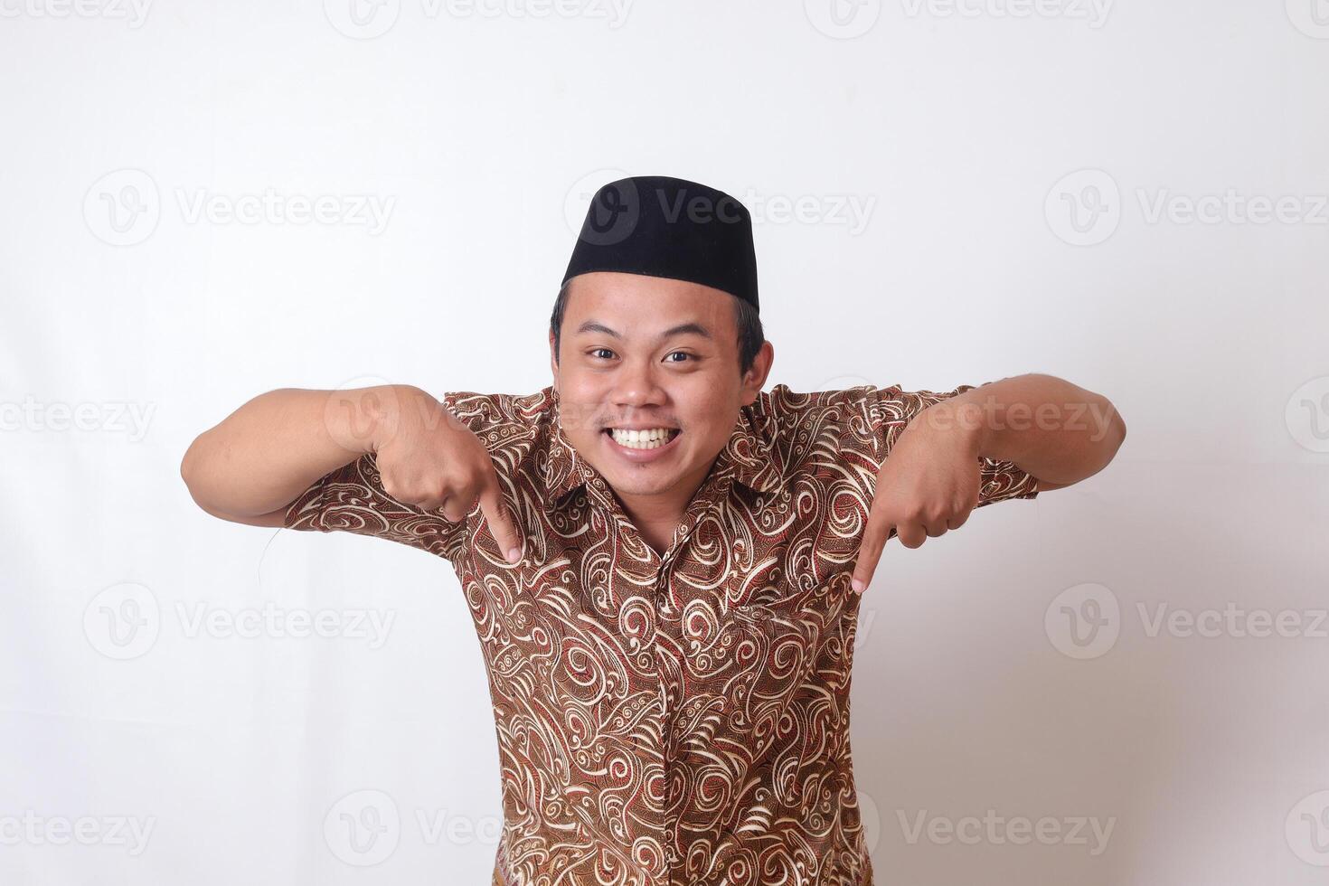 retrato do animado ásia homem vestindo batik camisa e Songkok sorridente e olhando às a Câmera apontando com dois mãos e dedos para a baixa lado. isolado imagem em cinzento fundo foto