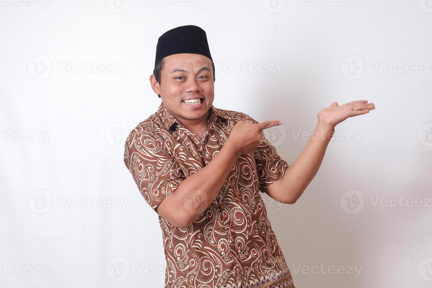 retrato do animado ásia homem vestindo batik camisa e Songkok sorridente e olhando às a Câmera apontando com dois mãos e dedos para a lado. isolado imagem em cinzento fundo foto