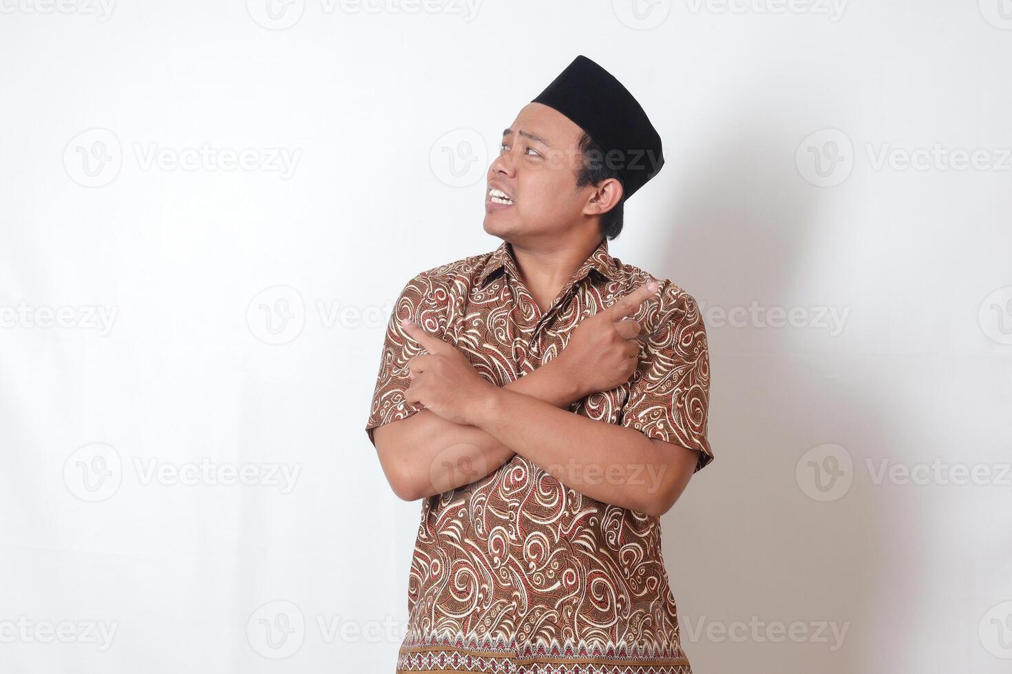 retrato do confuso ásia homem vestindo batik camisa e Songkok com cruzado mãos, apontando de lado, fazer escolha, escolhendo entre dois objetos. isolado imagem em cinzento fundo foto