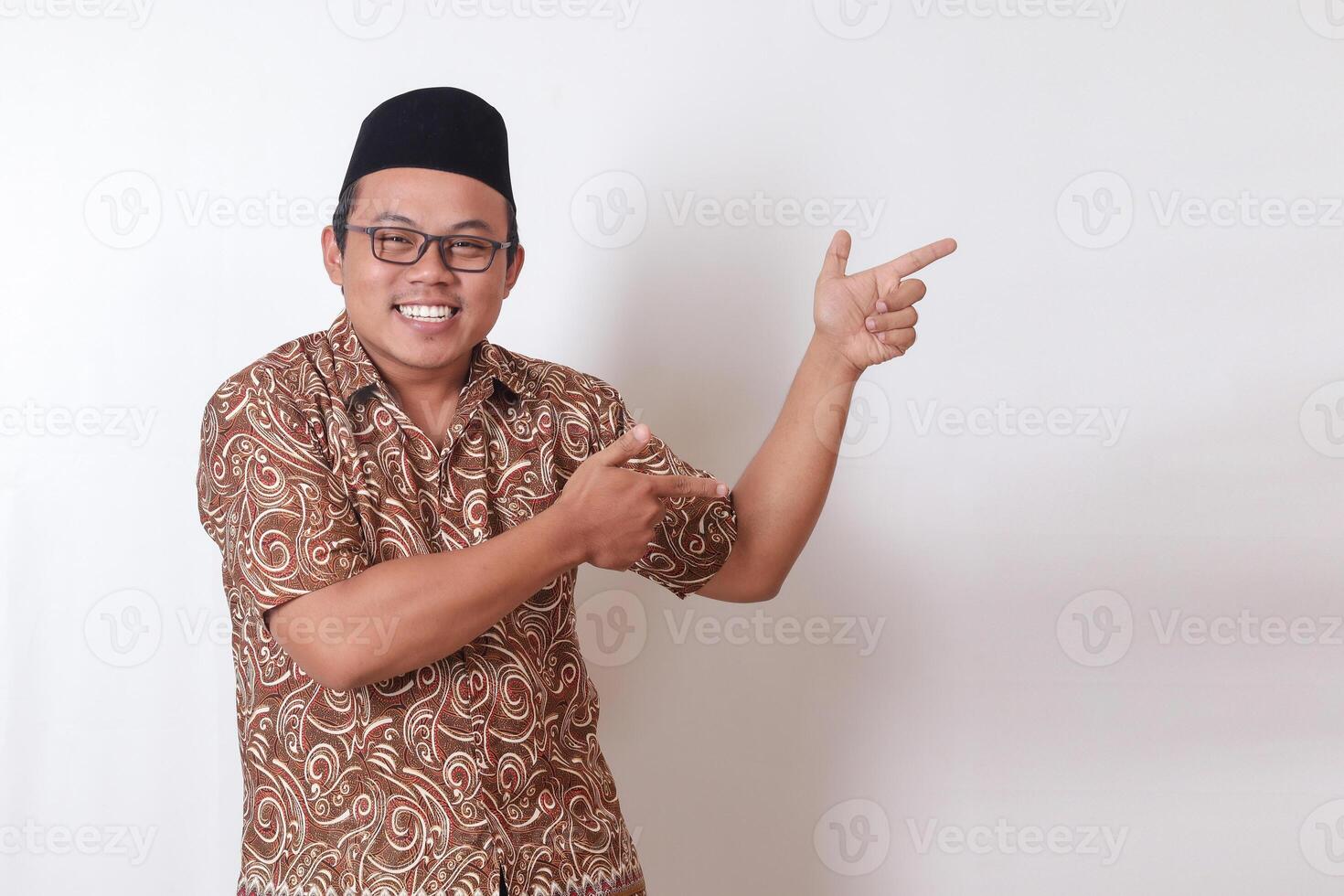 retrato do animado ásia homem vestindo batik camisa e Songkok sorridente e olhando às a Câmera apontando com dois mãos e dedos para a lado. isolado imagem em cinzento fundo foto